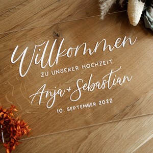 Op de afbeelding: Een helder acryl bord met witte letters met de tekst "Willkommen zu unserer Hochzeit Anja + Sebastian 10. September 2022".