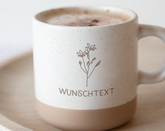 Könnte beinhalten: Eine weiße Keramiktasse mit gesprenkelter Oberfläche und beigem Boden, gefüllt mit Cappuccino. Die Tasse hat ein Blumendesign und das Wort "WUNSCHTEXT" in Braun. Die Tasse steht auf einem Holztablett.