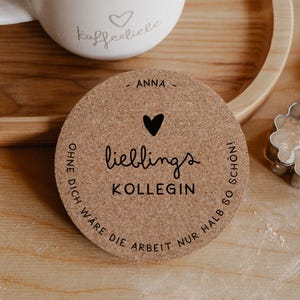 Könnte beinhalten: Ein runder Untersetzer aus Kork mit dem Text "lieblings KOLLEGIN" und einem schwarzen Herz. Der Untersetzer trägt auch den Namen "ANNA" und weiteren Text am Rand. Eine weiße Tasse mit dem Wort "kaffeeliebe" ist im Hintergrund.
