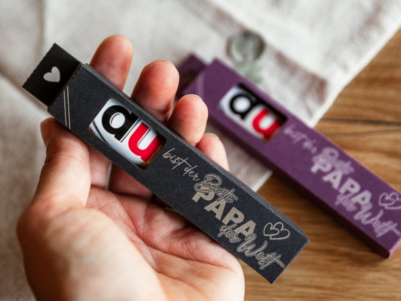 May include: Two small black boxes with the text "Du bist der beste Papa der Welt" and "Du bist der beste Papa der Welt" in gold lettering. The boxes are decorated with a heart and the word "Du" in red and white lettering.