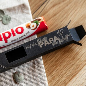 May include: A black gift box with a window to display a chocolate bar. The box has gold lettering that reads "bist der Beste PAPA der Welt" which translates to "You are the best Dad in the world".