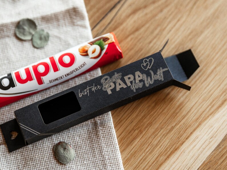 May include: A black gift box with a window to display a chocolate bar. The box has gold lettering that reads "bist der Beste PAPA der Welt" which translates to "You are the best Dad in the world".