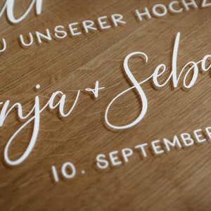 Op de afbeelding: Een wit acryl bord met de tekst "Anja + Sebastian" en de datum "10. September 202" voor een bruiloft.