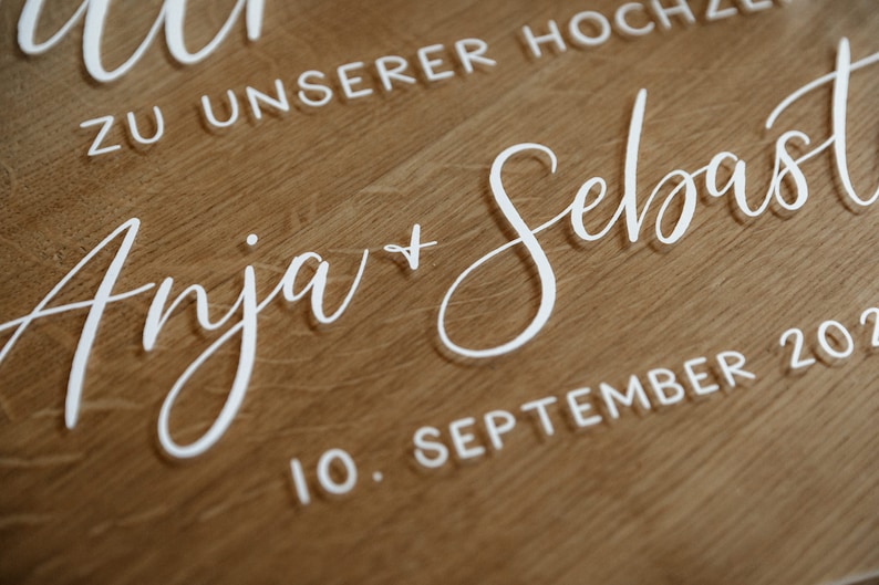 Op de afbeelding: Een wit acryl bord met de tekst "Anja + Sebastian" en de datum "10. September 202" voor een bruiloft.