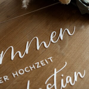 Op de afbeelding: Een helder acryl bord met de woorden "Willkommen" en "unserer Hochzeit" in wit cursief schrift.