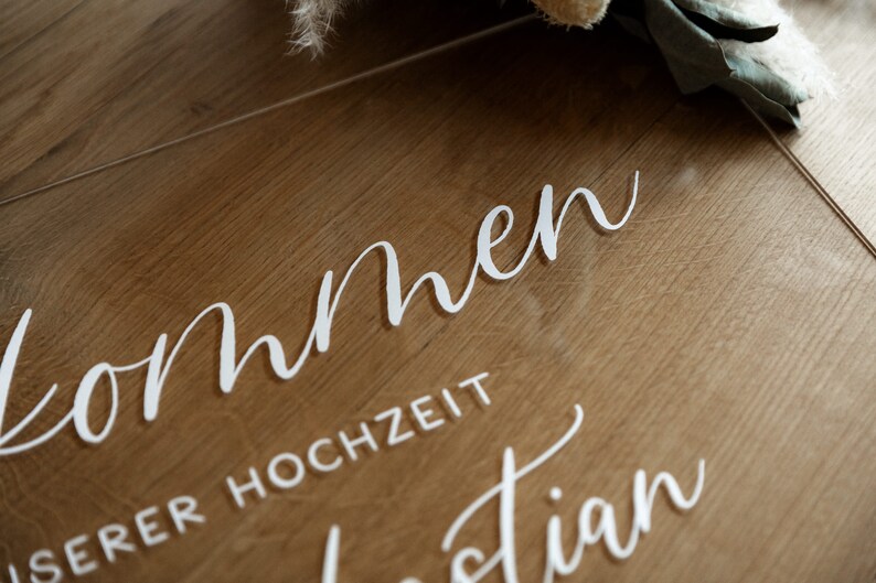 Op de afbeelding: Een helder acryl bord met de woorden "Willkommen" en "unserer Hochzeit" in wit cursief schrift.
