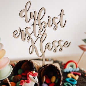 Peut inclure: Un gâteau au chocolat décoré de bonbons colorés, d'un arc-en-ciel et d'une figurine de licorne. Le gâteau est surmonté d'une pancarte en bois qui dit "Du bist erste Klasse".
