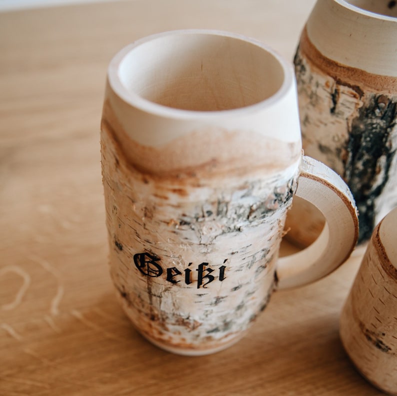 May include: A wooden mug with a birch bark exterior. The mug has a handle and the word "Gei&szlig;l" is engraved on the side.