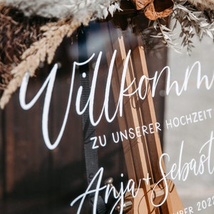 Op de afbeelding: Een helder acryl bord met de tekst "Willkommen zu unserer Hochzeit Anja + Sebastian" geschreven in witte kalligrafie. Het bord is versierd met gedroogde bloemen en groen.