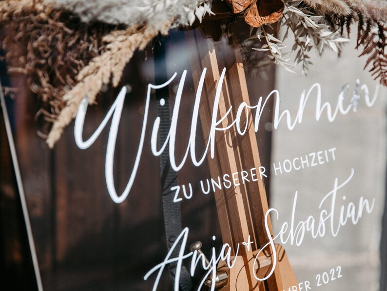 Op de afbeelding: Een helder acryl bord met de tekst "Willkommen zu unserer Hochzeit Anja + Sebastian" geschreven in witte kalligrafie. Het bord is versierd met gedroogde bloemen en groen.