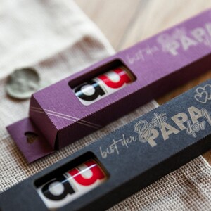 May include: Two small gift boxes with a heart-shaped cutout on the side. The boxes are purple and black and have a gold inscription that reads "Best Papa in the World".