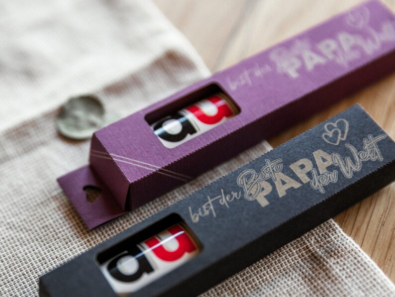 May include: Two small gift boxes with a heart-shaped cutout on the side. The boxes are purple and black and have a gold inscription that reads "Best Papa in the World".