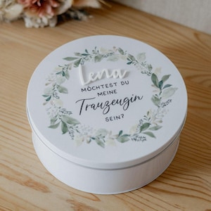 May include: A white, round gift box with a lid featuring the name "Lena" in raised letters. The lid is decorated with a green and white floral wreath and text that reads "Möchtest du meine Trauzeugin sein?"