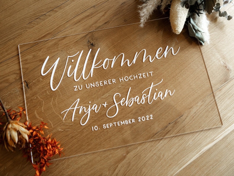 Op de afbeelding: Een helder acryl bord met witte letters met de tekst "Willkommen zu unserer Hochzeit Anja + Sebastian 10. September 2022".