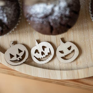 Könnte beinhalten: Drei Kürbis-Ausschnitte aus Holz mit geschnitzten Gesichtern liegen auf einem Holztablett, in der Nähe von zwei Muffins. Die Kürbisse haben unterschiedliche Ausdrücke, ideal für Halloween-Dekorationen. Das Tablett ist rund und aus hellem Holz.
