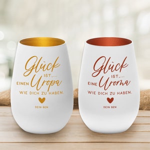 Könnte beinhalten: Zwei weiße Keramik-Weingläser mit goldenen und kupfernen Rändern. Die Gläser haben den Text "Glück ist... einen Uropa wie dich zu haben." und "Glück ist... eine Uroma wie dich zu haben." aufgedruckt. Der Text ist in einer kursivschriftlichen Schriftart und in der Farbe Gold auf dem linken Glas und Kupfer auf dem rechten Glas.  Es befindet sich ein kleines Herzsymbol unter dem Text auf jedem Glas. Der Text "Dein Ben" ist unter dem Herzen auf jedem Glas aufgedruckt.