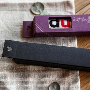 May include: Two small boxes, one purple and one black. The purple box has the text "du" in red and white letters and the text "bist der Beste Papier" in white letters. The black box has a heart-shaped cutout.