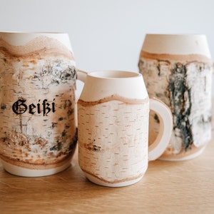 May include: Three birch bark mugs, two large and one small, with a natural wood grain pattern. The largest mug has the word "Geiki" inscribed on it.