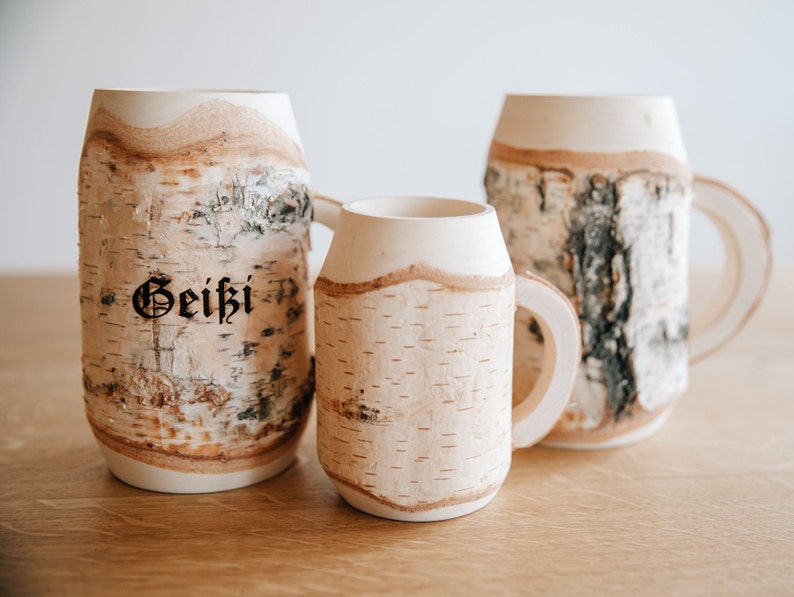 May include: Three birch bark mugs, two large and one small, with a natural wood grain pattern. The largest mug has the word "Geiki" inscribed on it.