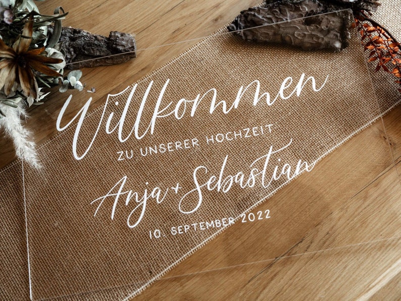 Op de afbeelding: Een helder acryl bord met witte kalligrafie waarop staat "Willkommen zu unserer Hochzeit Anja + Sebastian 10. September 2022". Het bord ligt op een jute oppervlak.