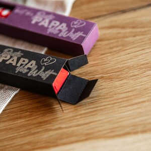 May include: Two small black and purple boxes with the text "Beste Papa der Welt" (Best Dad in the World) and two hearts on them. The boxes are open and show a red lip balm inside.