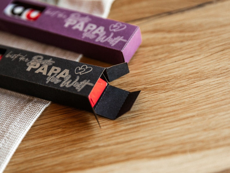 May include: Two small black and purple boxes with the text "Beste Papa der Welt" (Best Dad in the World) and two hearts on them. The boxes are open and show a red lip balm inside.