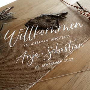 Op de afbeelding: Duidelijk acryl huwelijksbord met de tekst "Willkommen zu unserer Hochzeit Anja & Sebastian 10. September 2022" in witte kalligrafie.