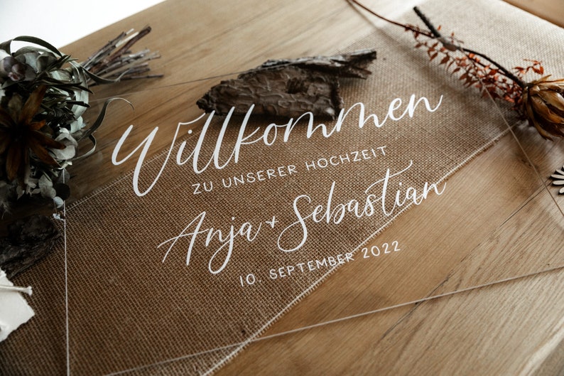 Op de afbeelding: Duidelijk acryl huwelijksbord met de tekst "Willkommen zu unserer Hochzeit Anja & Sebastian 10. September 2022" in witte kalligrafie.