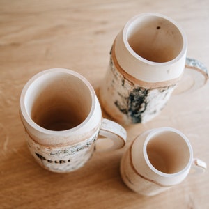 May include: Three wooden mugs with a birch bark design. The mugs are all different sizes and have a natural wood finish.