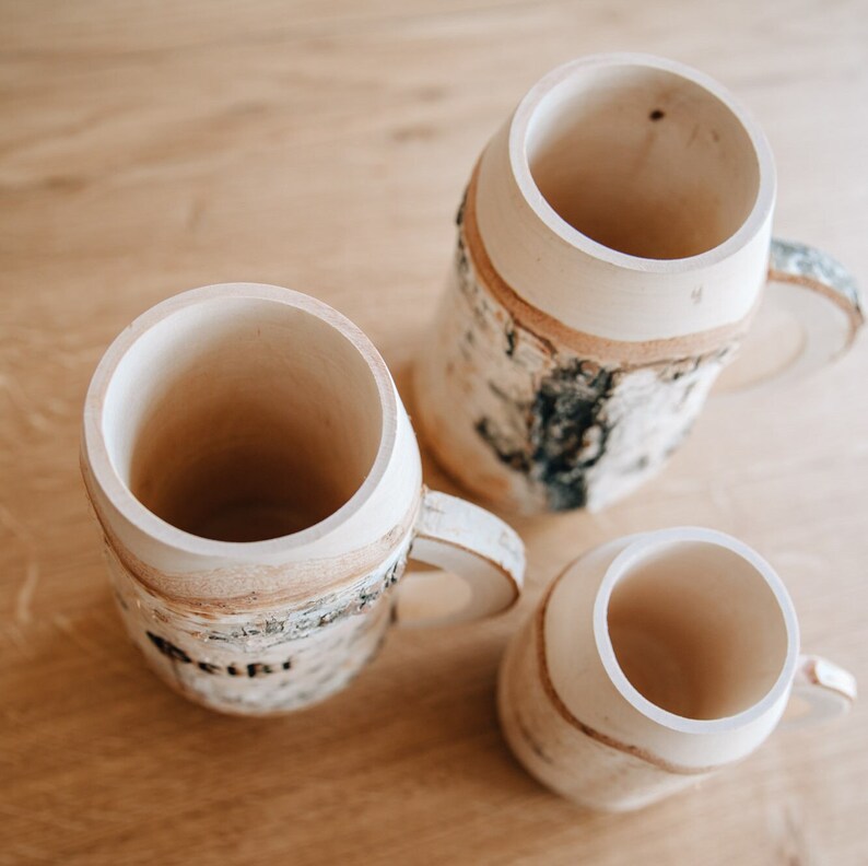 May include: Three wooden mugs with a birch bark design. The mugs are all different sizes and have a natural wood finish.