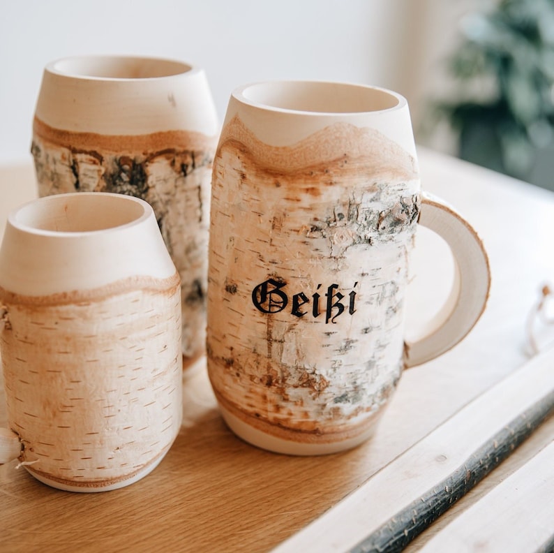 May include: Three wooden mugs made from birch bark. The mug on the right has the word "Geiki" written on it in black.