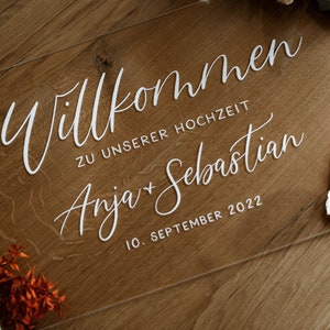 Op de afbeelding: Een helder acryl bord met witte kalligrafie waarop staat "Willkommen zu unserer Hochzeit Anja + Sebastian 10. September 2022".