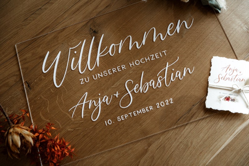 Op de afbeelding: Een helder acryl bord met witte kalligrafie waarop staat "Willkommen zu unserer Hochzeit Anja + Sebastian 10. September 2022".