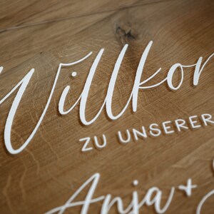 Op de afbeelding: Een wit acryl bord met de tekst "Willkommen zu unserer Hochzeit Anja + Seb" op een houten achtergrond.