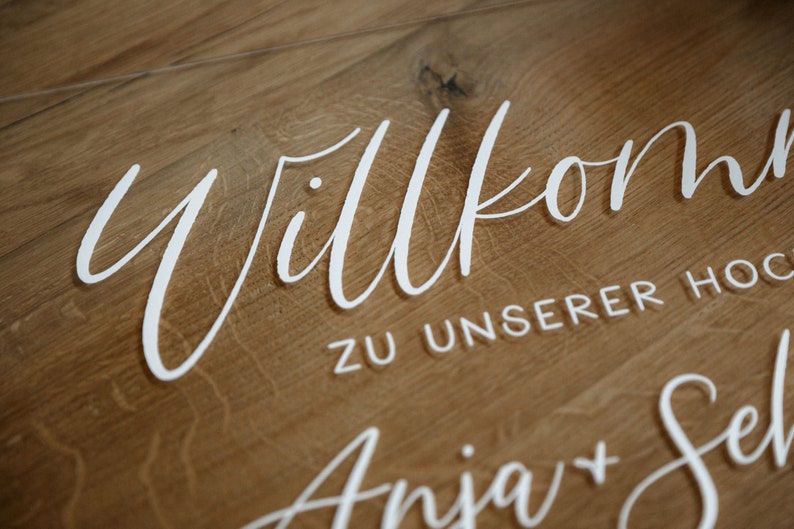 Op de afbeelding: Een wit acryl bord met de tekst "Willkommen zu unserer Hochzeit Anja + Seb" op een houten achtergrond.