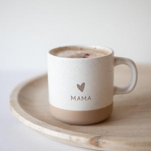 Op de afbeelding: Witte keramische mok met een gespikkelde afwerking en een bruine rand. De mok heeft een hartje en het woord "MAMA" erop gedrukt. De mok is gevuld met koffie en staat op een houten dienblad.