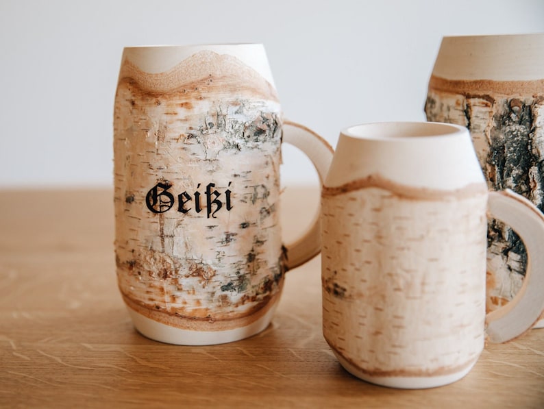 May include: Three white ceramic mugs with a birch bark design. The mug on the left has the word "Geiki" written on it in black.
