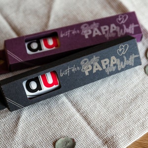 May include: Two black and purple boxes with a white background and gold lettering. The boxes have a small window showing a white card with the letters "au" in black and red. The boxes have the text "bist der Beste PAPA der Welt" on them.