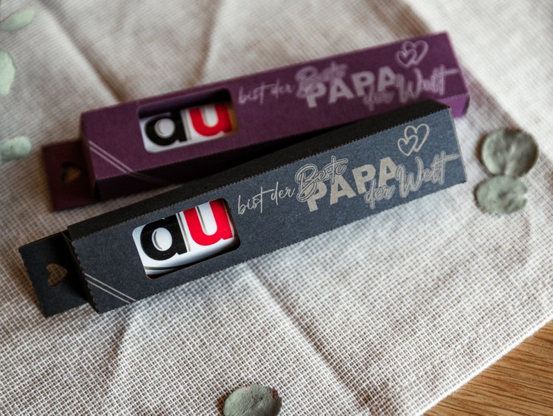 May include: Two black and purple boxes with a white background and gold lettering. The boxes have a small window showing a white card with the letters "au" in black and red. The boxes have the text "bist der Beste PAPA der Welt" on them.