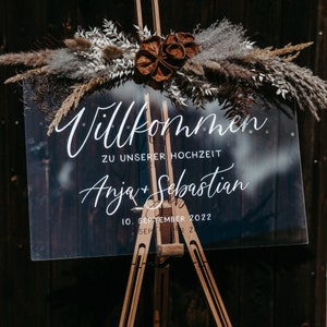 Op de afbeelding: Een helder acryl bord met witte kalligrafie waarop "Willkommen zu unserer Hochzeit Anja + Sebastian 10. September 2022" staat, is op een houten ezel geplaatst. Het bord is versierd met gedroogd pampasgras en bruine bloemknoppen.