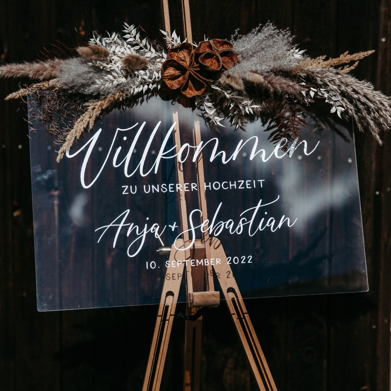 Op de afbeelding: Een helder acryl bord met witte kalligrafie waarop "Willkommen zu unserer Hochzeit Anja + Sebastian 10. September 2022" staat, is op een houten ezel geplaatst. Het bord is versierd met gedroogd pampasgras en bruine bloemknoppen.