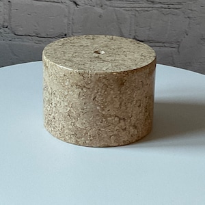 May include: A round, beige marble pedestal with a small hole in the centre. The pedestal has a speckled pattern and is sitting on a white surface.