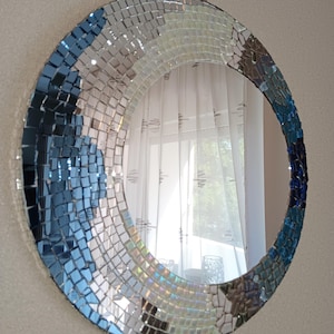 Könnte beinhalten: Runder Mosaikspiegel mit einem Rand aus kleinen, quadratischen Fliesen in Blau-, Weiß- und Silbertönen. Der Spiegel wird an einer Kordel aus Naturfaser aufgehängt. Das Mosaikdesign erzeugt einen strukturierten, künstlerischen Look.