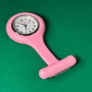 Op de afbeelding: Een roze quartz verpleegster zakhorloge met een witte wijzerplaat en zwarte cijfers. Het horloge heeft een speld om te bevestigen en een stift om op te winden. Het horloge staat tegen een groene achtergrond.