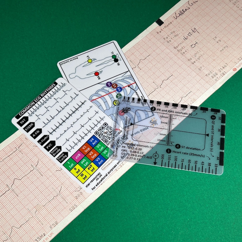 May include: A clear plastic ruler with markings for measuring heart rate, PR, QRS, QT, and ST intervals. The ruler is labeled with "Expected Intervals (s)" and includes measurements in millimeters. The ruler is lying on top of a white card with a diagram of the human body and a chart of common ECG rhythms.
