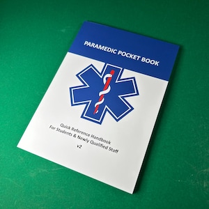 May include: A paramedic pocket book with a blue and white cover. The cover features a blue Star of Life emblem with a red staff and white snake. Text on the cover reads "PARAMEDIC POCKET BOOK" and "Quick Reference Handbook For Students & Newly Qualified Staff v2."