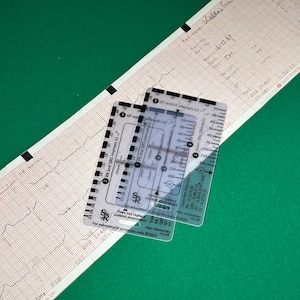 May include: A clear plastic ruler with markings for measuring heart rate, PR, QRS, and QT intervals. The ruler is labeled "Star-resources" and includes expected intervals for PR, QT, QRS, and ST. The ruler is on top of a white electrocardiogram printout.