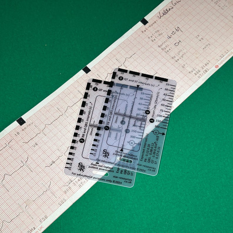 May include: A clear plastic ruler with markings for measuring heart rate, PR, QRS, and QT intervals. The ruler is labeled "Star-resources" and includes expected intervals for PR, QT, QRS, and ST. The ruler is on top of a white electrocardiogram printout.