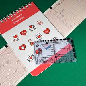 May include: A white plastic ECG ruler with red and blue markings. The ruler has a scale for measuring PR and QRS intervals, ST deviation, and heart rate. The ruler also includes expected intervals for PR, QRS, QT, and ST segments. The ruler is lying on a green surface next to a red and white notebook with ECG pocket flash cards.