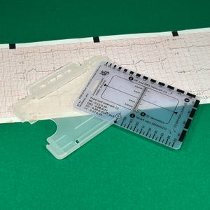 May include: A clear plastic ruler with markings for measuring heart rate, ST deviation, and PR and QRS intervals in seconds. The ruler is labeled with expected intervals for PR, QRS, QT, and ST segments. The ruler is on a green surface.